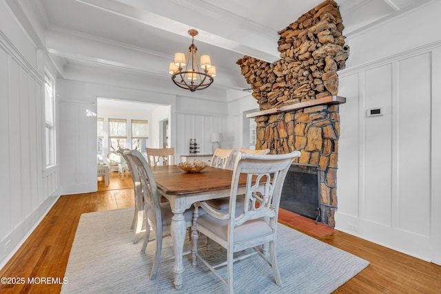 dining room with a notable chandelier, beam ceiling, crown molding, light wood-type flooring, and a stone fireplace