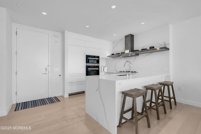 kitchen with kitchen peninsula, a kitchen breakfast bar, wall chimney range hood, light hardwood / wood-style flooring, and white cabinets