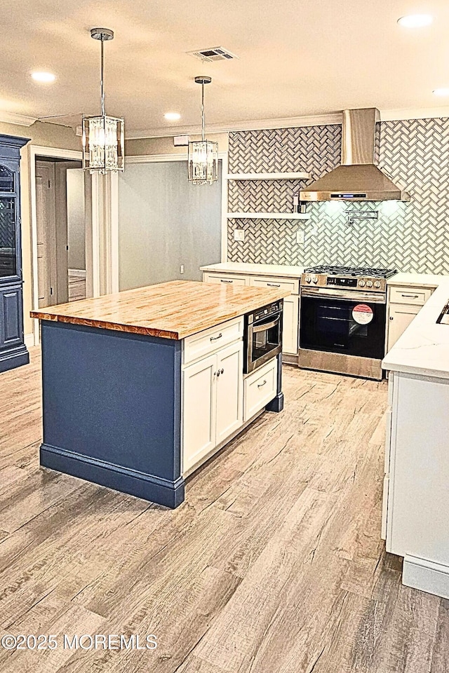 kitchen with stainless steel range with gas stovetop, hanging light fixtures, wall chimney exhaust hood, white cabinets, and butcher block counters