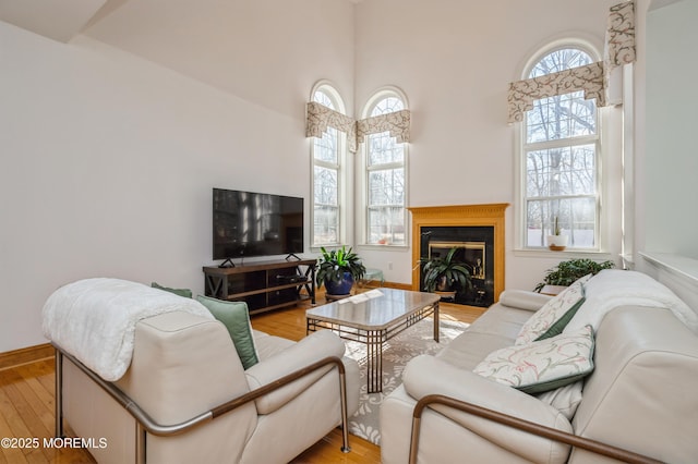 living room with light wood-type flooring, a towering ceiling, baseboards, and a glass covered fireplace