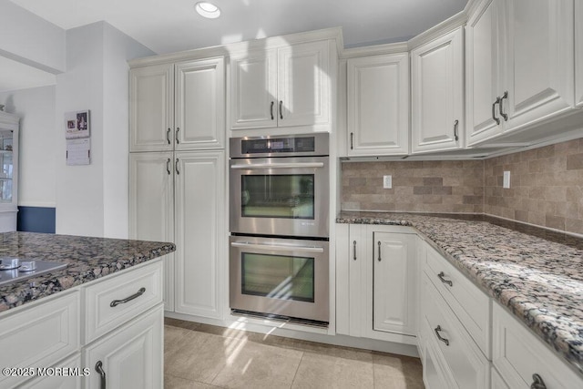 kitchen featuring light tile patterned floors, stainless steel double oven, backsplash, dark stone counters, and white cabinets