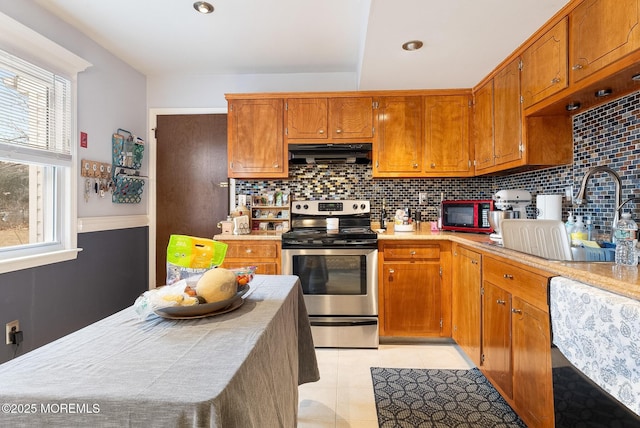 kitchen with brown cabinets, stainless steel range with electric cooktop, under cabinet range hood, backsplash, and light tile patterned flooring