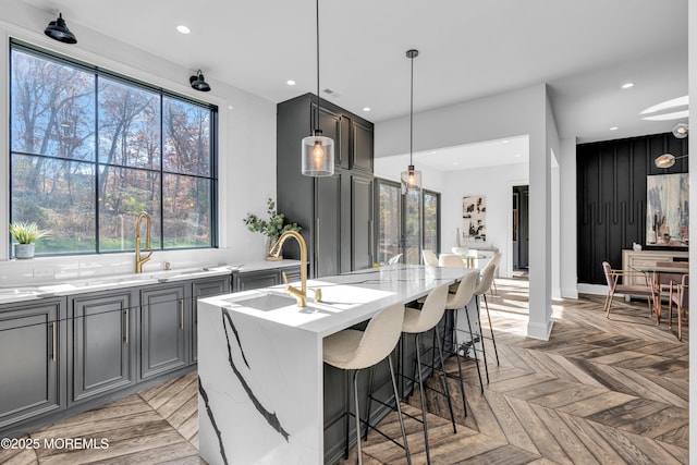 kitchen featuring light parquet floors, a center island with sink, gray cabinetry, light stone countertops, and pendant lighting