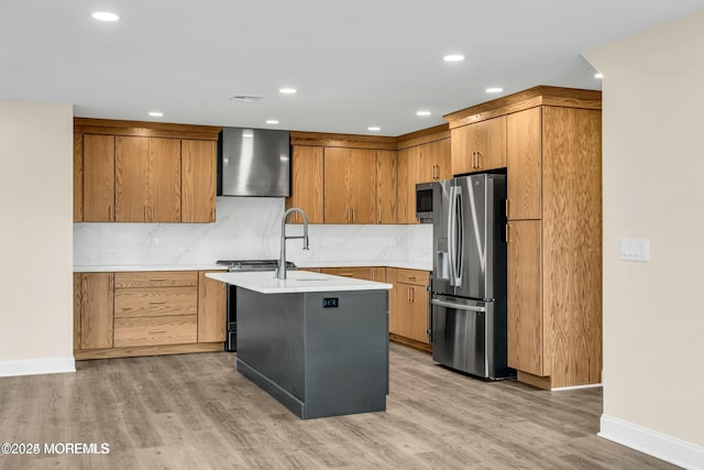 kitchen featuring a kitchen island with sink, appliances with stainless steel finishes, light hardwood / wood-style floors, wall chimney range hood, and tasteful backsplash