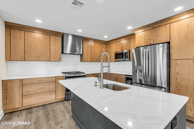 kitchen featuring wall chimney exhaust hood, light wood-type flooring, a kitchen island with sink, appliances with stainless steel finishes, and sink