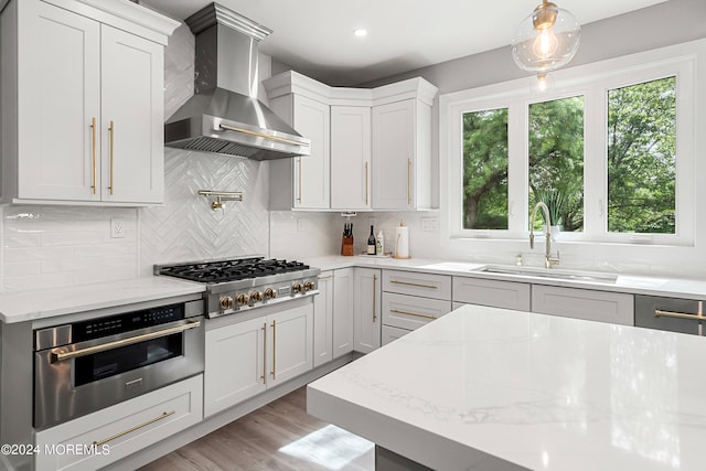 kitchen featuring wall chimney range hood, stainless steel appliances, sink, backsplash, and light stone counters