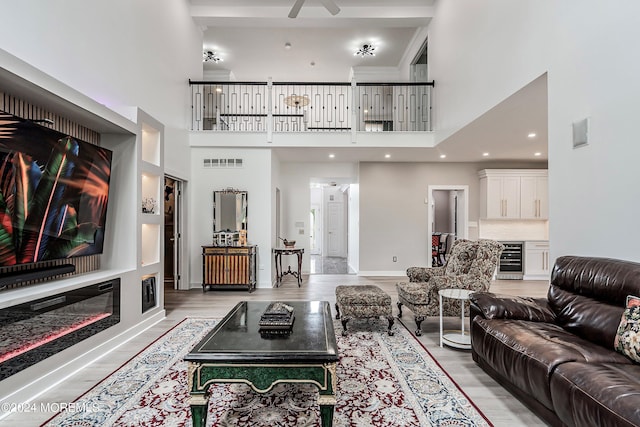 living room with ceiling fan, light wood-type flooring, a towering ceiling, and beverage cooler