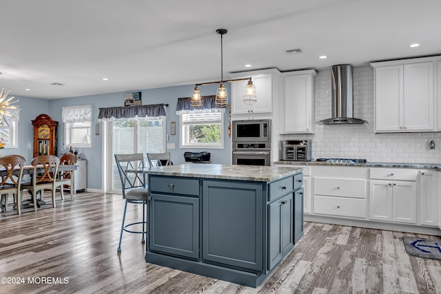 kitchen with white cabinets, a center island, wall chimney exhaust hood, appliances with stainless steel finishes, and light wood-type flooring