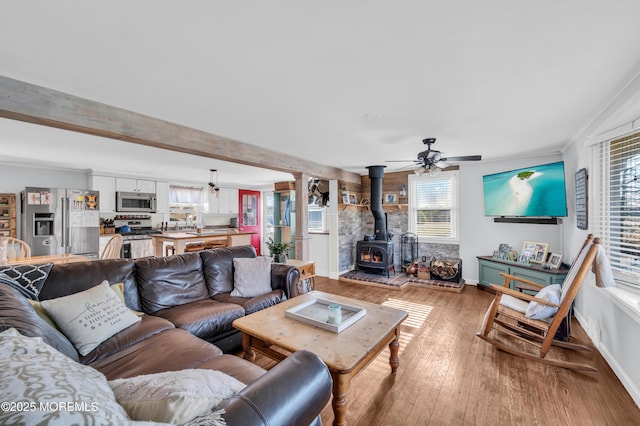 living room featuring beamed ceiling, a wood stove, light wood-type flooring, ceiling fan, and crown molding