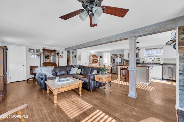 living room with ceiling fan, light hardwood / wood-style floors, beam ceiling, sink, and decorative columns