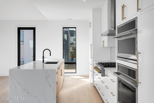 kitchen with white cabinets, wall chimney exhaust hood, an island with sink, sink, and light stone counters