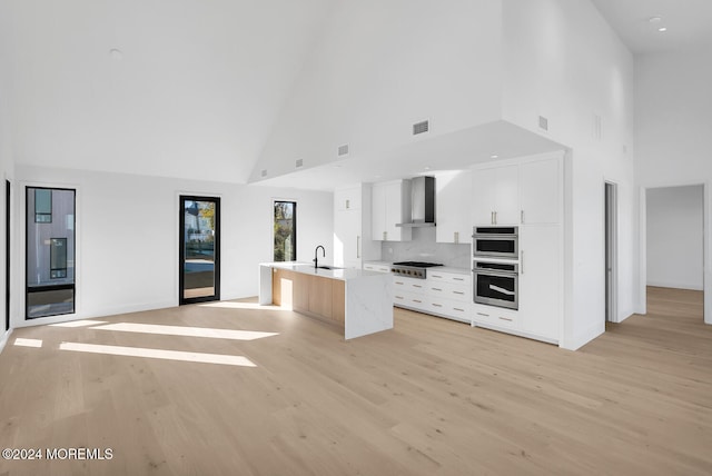 kitchen with a kitchen island with sink, white cabinets, wall chimney range hood, high vaulted ceiling, and backsplash