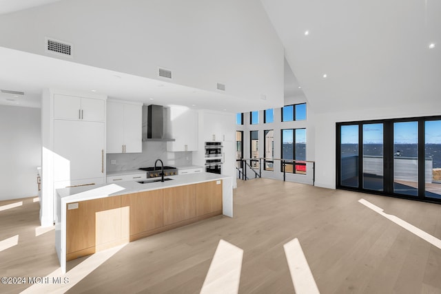 kitchen featuring a large island with sink, wall chimney exhaust hood, white cabinetry, tasteful backsplash, and high vaulted ceiling