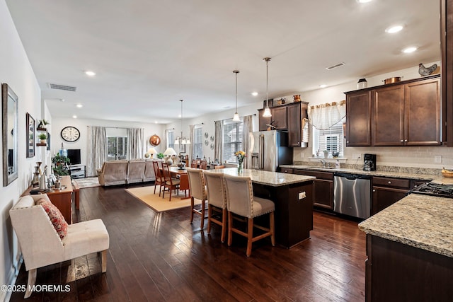 kitchen with appliances with stainless steel finishes, hanging light fixtures, a kitchen island, dark wood-type flooring, and light stone counters