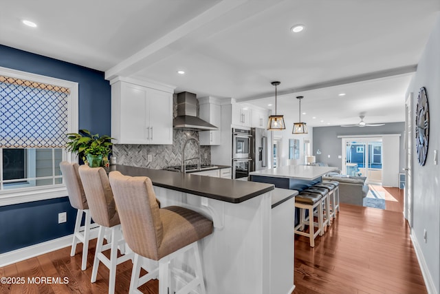 kitchen with wall chimney range hood, hanging light fixtures, white cabinets, a kitchen bar, and dark hardwood / wood-style flooring