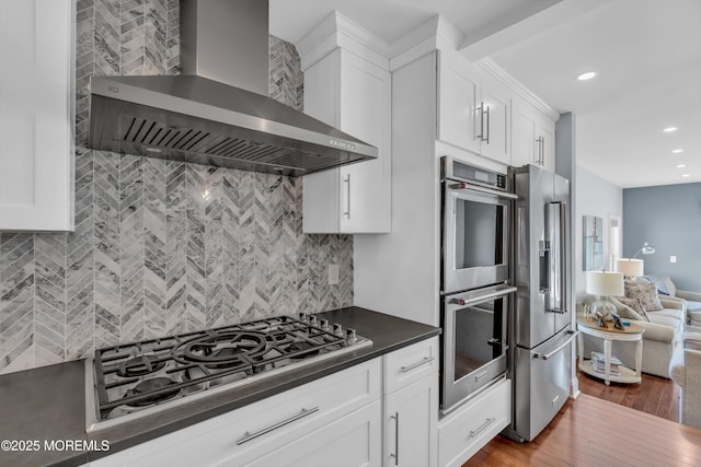 kitchen with wall chimney range hood, wood-type flooring, white cabinets, and appliances with stainless steel finishes