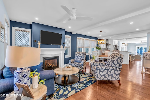 living room with ceiling fan, wood-type flooring, a premium fireplace, and beam ceiling