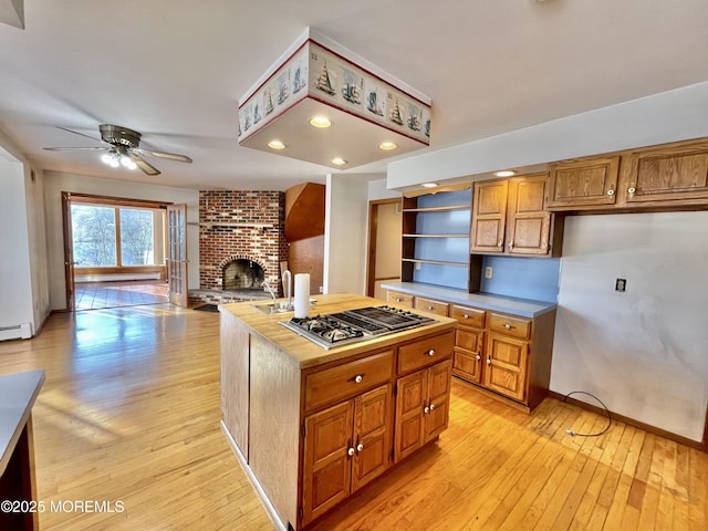 kitchen with ceiling fan, a center island, light hardwood / wood-style floors, stainless steel gas cooktop, and a brick fireplace