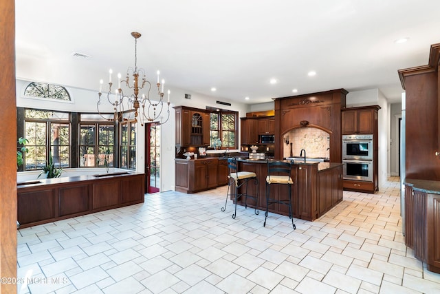 kitchen with pendant lighting, appliances with stainless steel finishes, an island with sink, decorative backsplash, and a chandelier