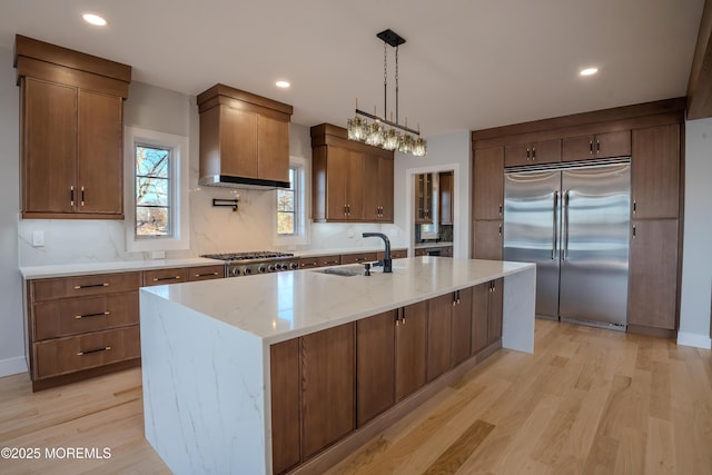 kitchen featuring sink, hanging light fixtures, stainless steel appliances, custom range hood, and a center island with sink