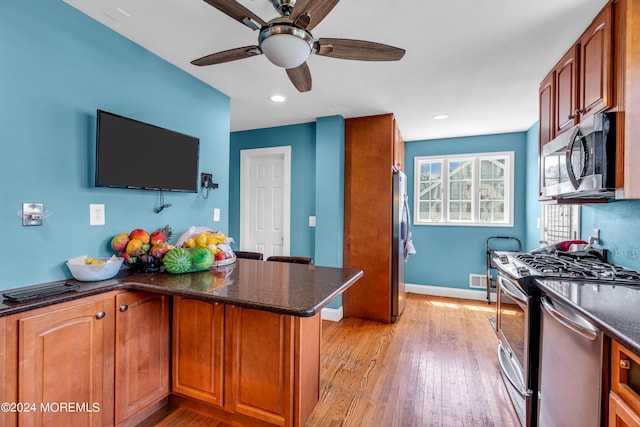 kitchen with ceiling fan, appliances with stainless steel finishes, kitchen peninsula, dark stone counters, and light wood-type flooring