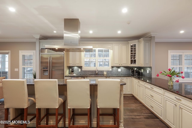 kitchen featuring island exhaust hood, built in refrigerator, a kitchen breakfast bar, and dark stone countertops