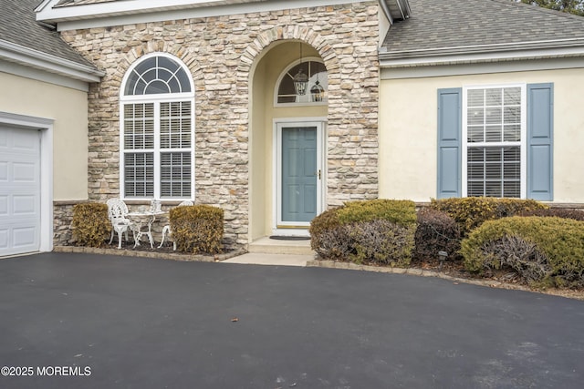 view of exterior entry featuring a garage, stone siding, roof with shingles, and stucco siding