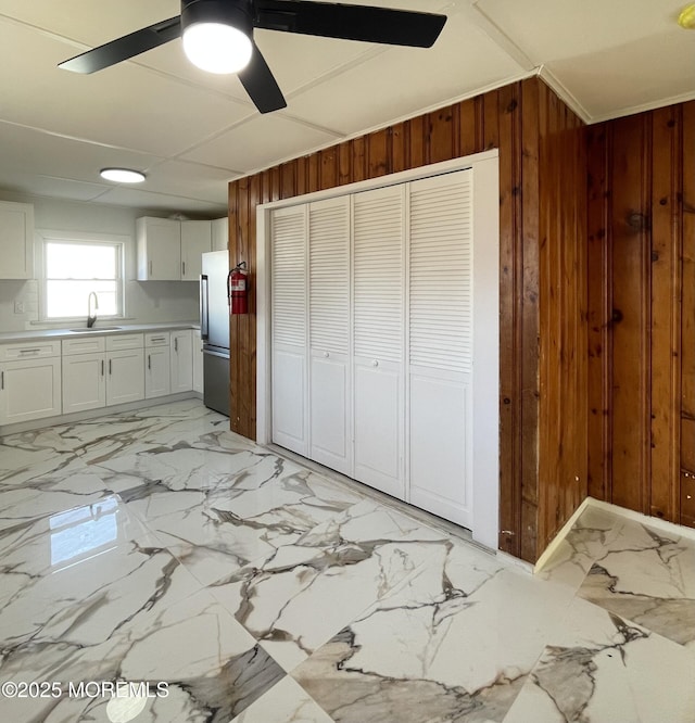 kitchen featuring sink, wood walls, white cabinetry, stainless steel refrigerator, and ceiling fan