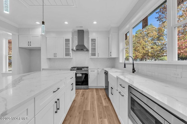 kitchen featuring wall chimney exhaust hood, white cabinetry, light stone counters, appliances with stainless steel finishes, and pendant lighting