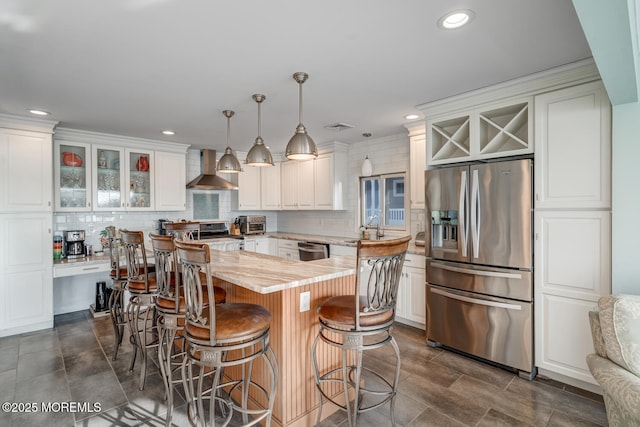 kitchen featuring pendant lighting, wall chimney range hood, appliances with stainless steel finishes, white cabinets, and a kitchen island