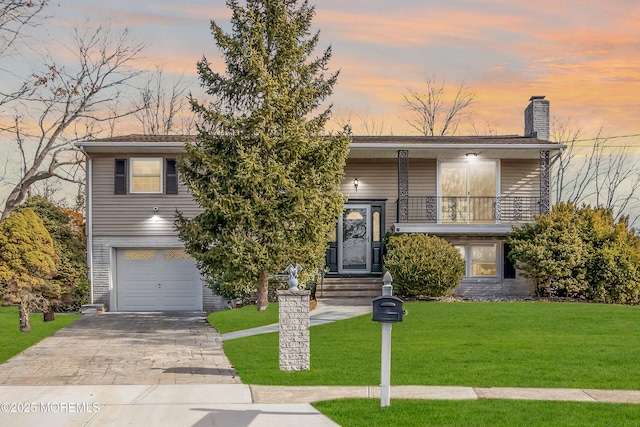 raised ranch with a balcony, a garage, decorative driveway, a lawn, and a chimney