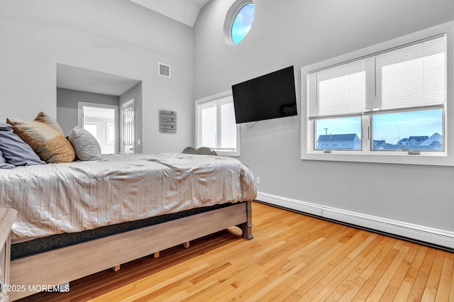 bedroom with a baseboard heating unit, hardwood / wood-style flooring, and a towering ceiling