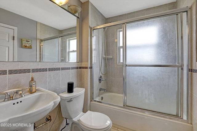 full bath featuring toilet, shower / bath combination with glass door, tile walls, and a sink