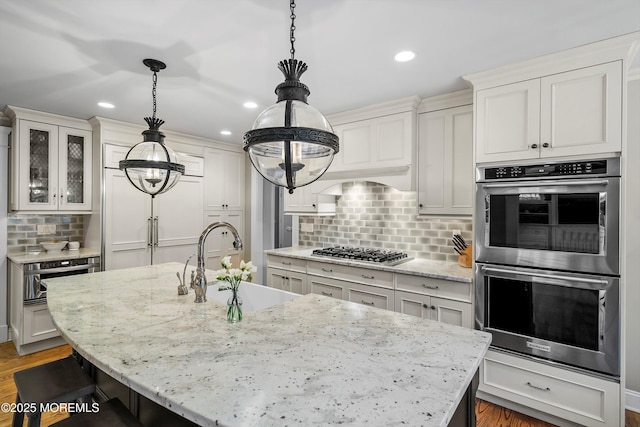 kitchen featuring an island with sink, backsplash, hanging light fixtures, stainless steel appliances, and light stone countertops