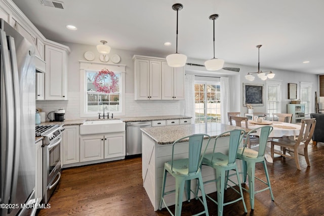 kitchen with appliances with stainless steel finishes, hanging light fixtures, light stone counters, white cabinets, and a kitchen island