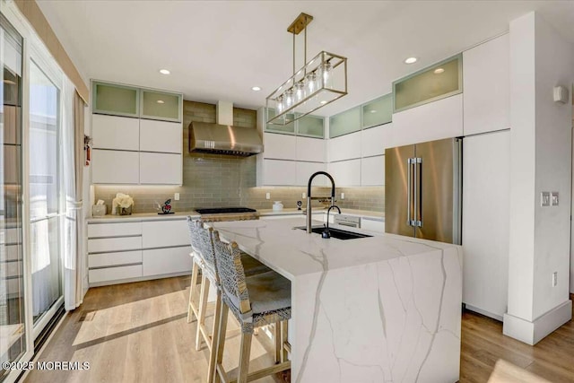 kitchen featuring pendant lighting, an island with sink, sink, white cabinets, and wall chimney range hood