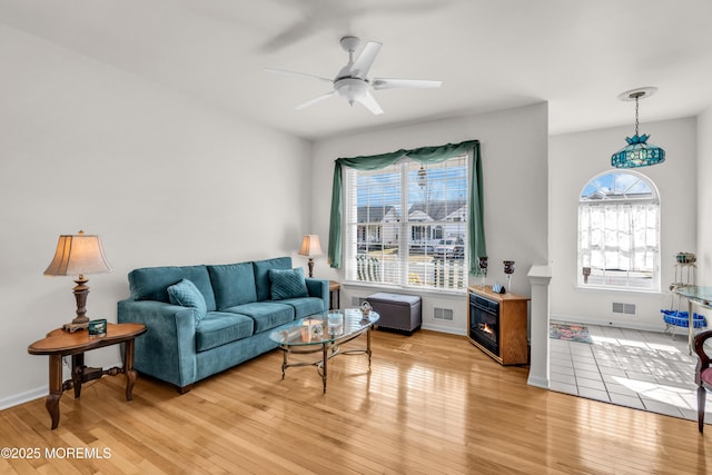 living room with ceiling fan, visible vents, baseboards, light wood-type flooring, and a glass covered fireplace