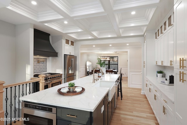 kitchen featuring high end appliances, custom range hood, glass insert cabinets, a kitchen island with sink, and white cabinetry