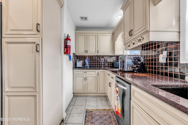 kitchen with light tile patterned flooring, decorative backsplash, dark stone counters, stainless steel dishwasher, and cream cabinetry