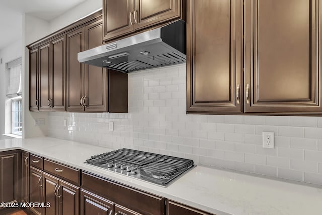 kitchen featuring dark brown cabinetry, light stone counters, tasteful backsplash, stainless steel gas cooktop, and exhaust hood