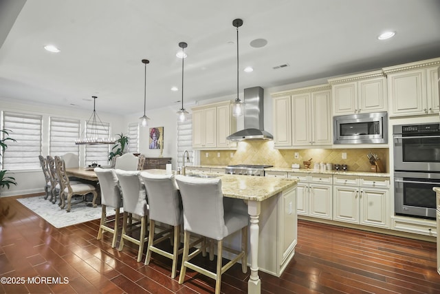 kitchen with stainless steel appliances, cream cabinets, a kitchen island with sink, and wall chimney exhaust hood