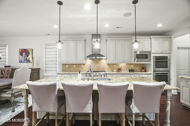 kitchen featuring appliances with stainless steel finishes, decorative light fixtures, light stone countertops, a center island with sink, and wall chimney exhaust hood