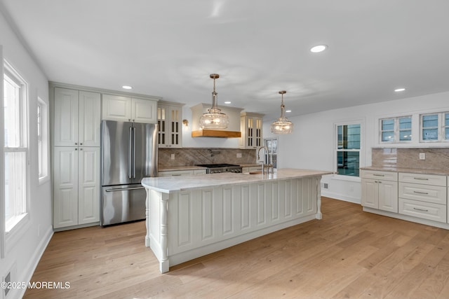 kitchen with stainless steel refrigerator, an island with sink, hanging light fixtures, light hardwood / wood-style floors, and light stone countertops