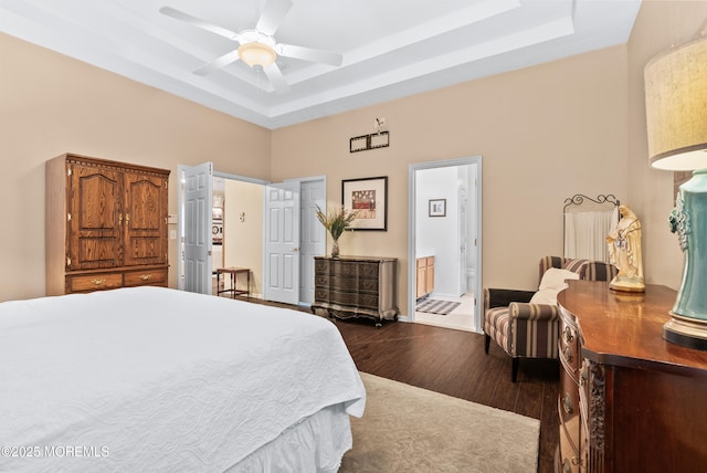 bedroom featuring a tray ceiling, dark wood-style flooring, a ceiling fan, ensuite bath, and baseboards