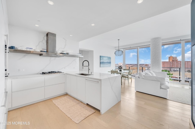 kitchen with kitchen peninsula, sink, white cabinets, wall chimney range hood, and pendant lighting