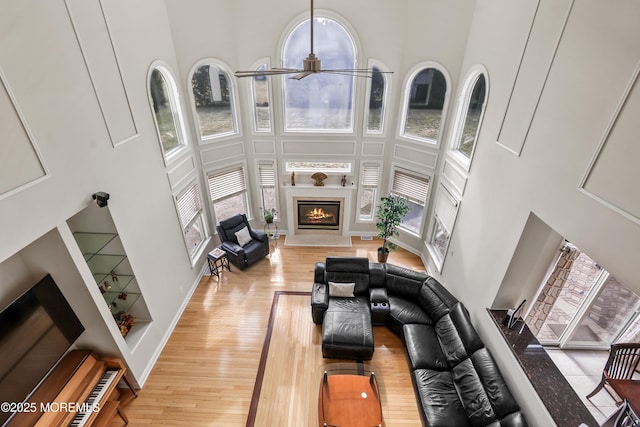 living room featuring a warm lit fireplace, ceiling fan, light wood-style flooring, a towering ceiling, and baseboards