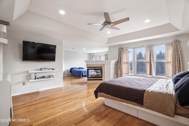 bedroom featuring visible vents, a raised ceiling, light wood-style floors, a multi sided fireplace, and recessed lighting