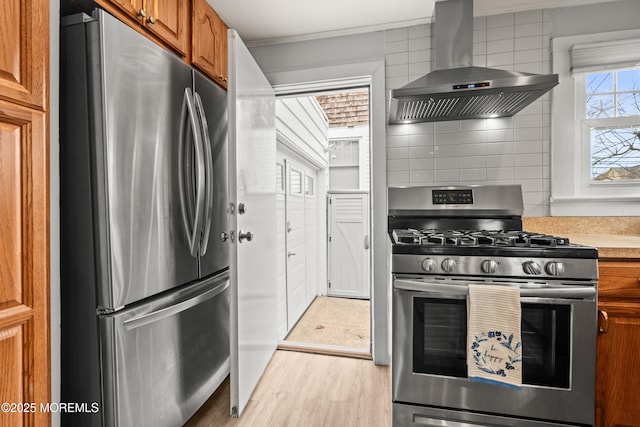 kitchen featuring decorative backsplash, light countertops, appliances with stainless steel finishes, wall chimney range hood, and brown cabinets