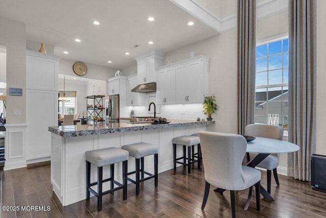 kitchen with under cabinet range hood, backsplash, a peninsula, a breakfast bar area, and stainless steel fridge with ice dispenser