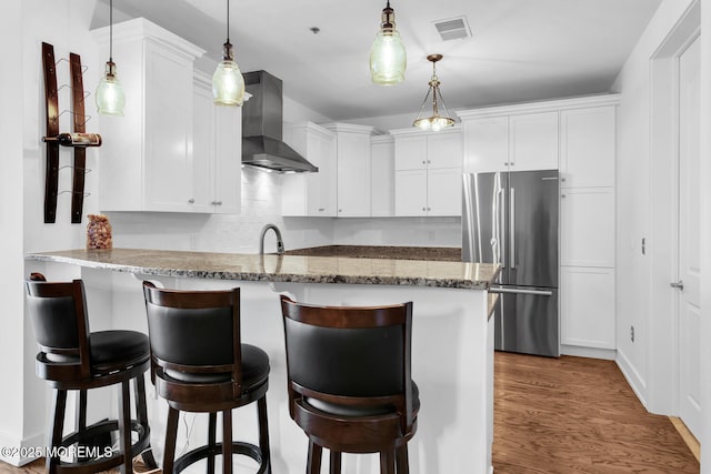 kitchen featuring white cabinetry, wall chimney range hood, stainless steel fridge, and kitchen peninsula